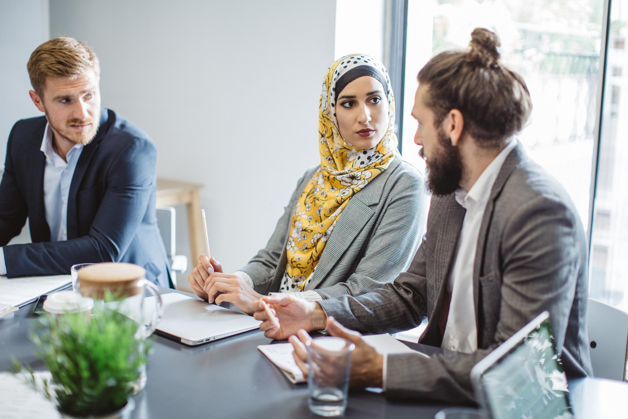 To menn og en kvinne sitter og diskuterer i et møte. Alle er kledd formelt, den ene mannen har kort velstelt hår, den andre har langt hår og skjegg. Kvinnen har hijab. 