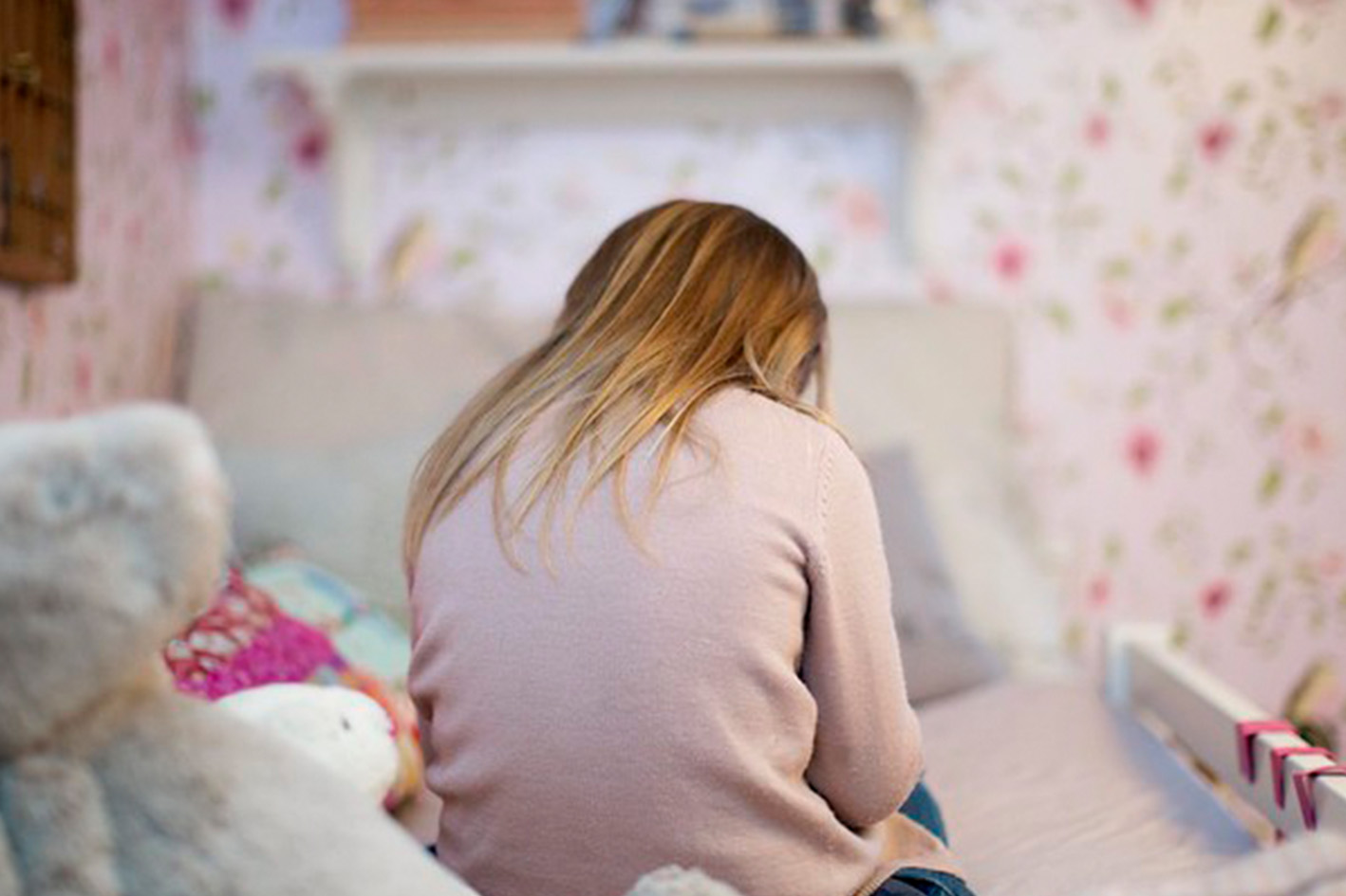 En ung jente i rosa genser og langt hår sitter vendt fra oss på senga. Hun har hodet bøyd