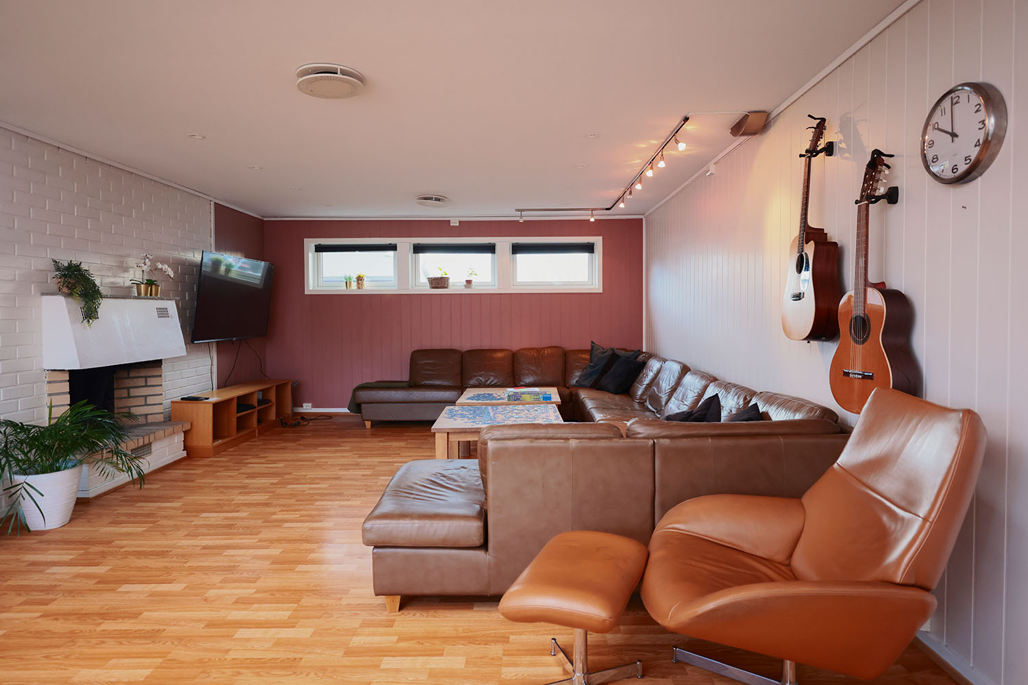 Stue med hjørnesofa, lenestol, tv, peis og gitarer på veggen. Foto.