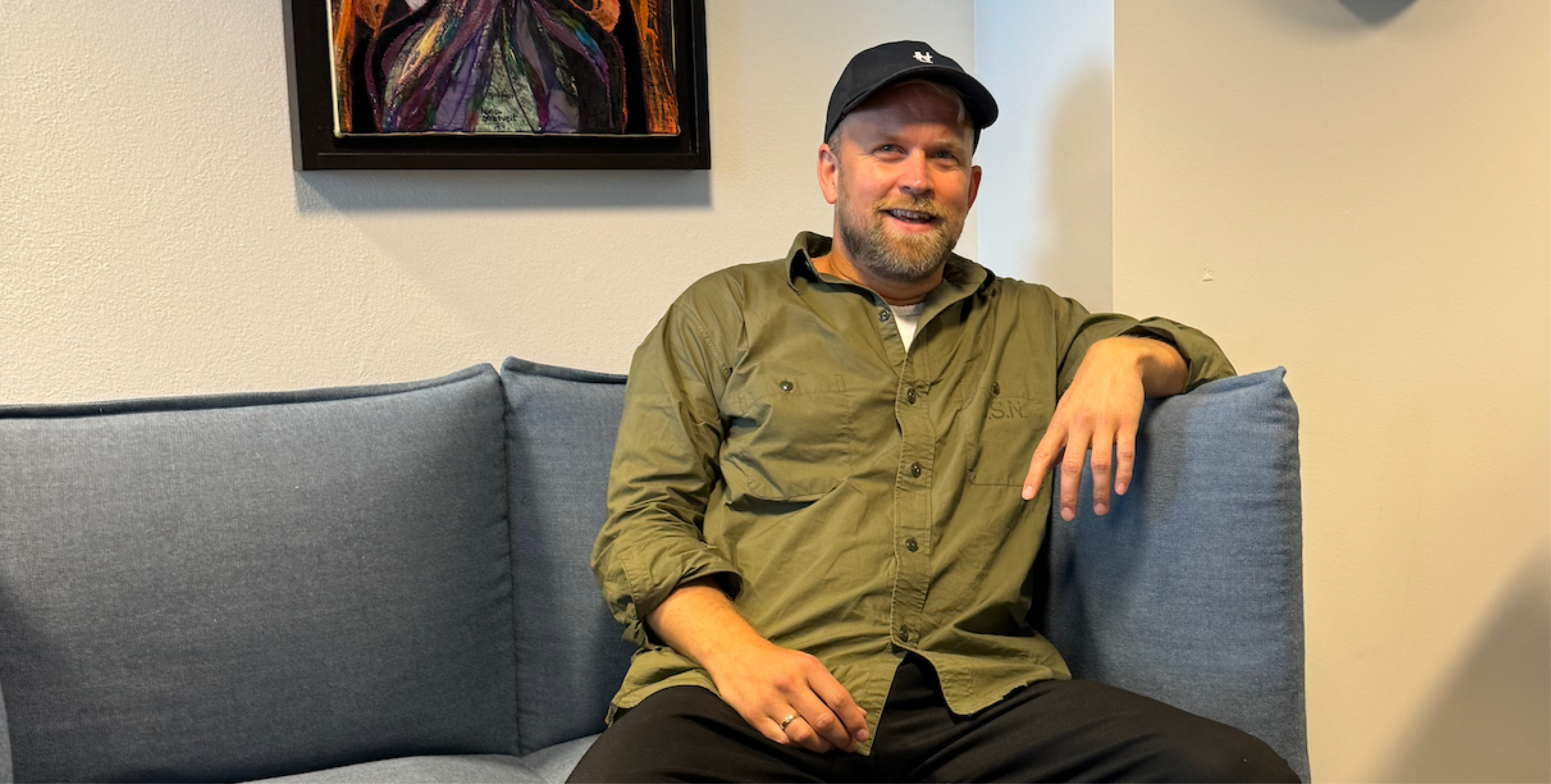 Portrett av mann med caps og skjorte som sitter i sofa på et kontor og smiler