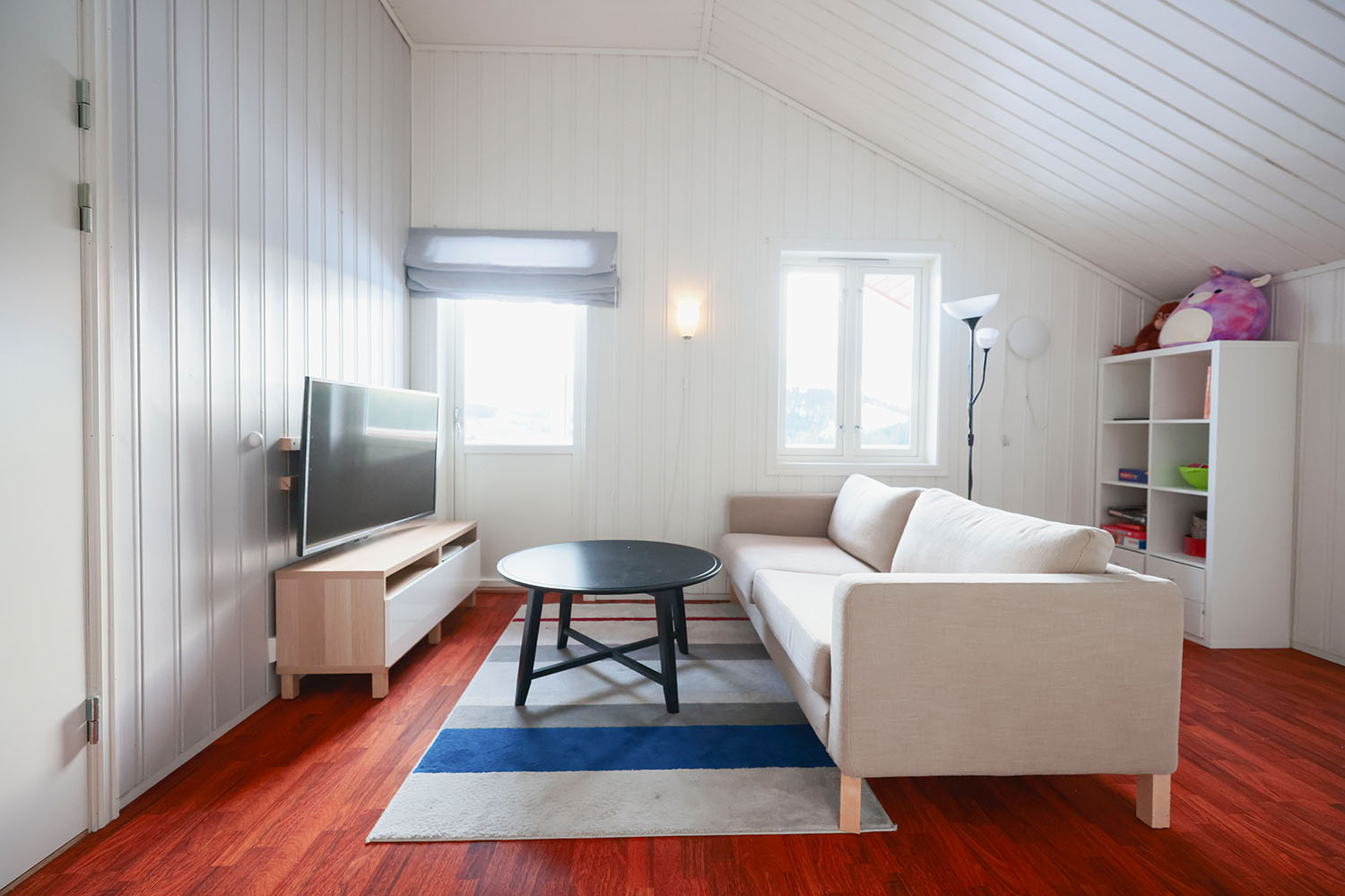 Loftsstue med sofa, bord, bokhylle og tv på veggen. Foto.